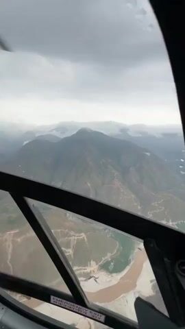 The video captures an aerial view from inside a helicopter or small aircraft, showcasing a dramatic mountainous landscape under an overcast sky. The camera looks out through the aircraft's windows, revealing rugged, green-tinged mountains with visible terracing or paths, a broad, muddy-brown river winding through a deep valley, and significant cloud cover clinging to the mountain peaks.

In the initial frames (0:00-0:01), the view is from a front-facing window, possibly part of the cockpit, showing a mountain range directly ahead and the river below. The text "EMERGENCY EXIT" is clearly visible on the lower frame of this window, along with partial numbers or letters, appearing to be "31". There appear to be raindrops or condensation on the window.

The camera then pans slightly to the right (0:01-0:02), revealing more of the internal structure of the aircraft, including a black handle on the window frame.

The final frames (0:02-0:04) offer a broader perspective, likely from a side window. The landscape is dominated by towering mountains, some partially obscured by thick white clouds. The wide, meandering river is prominently featured, cutting through the valley floor. The sky is entirely covered in dense, grey clouds, indicating an overcast day. The overall scene depicts a flight over a remote and majestic mountainous region, consistent with the provided location context of Rampur, Nepal.
