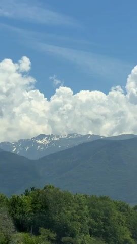 The video shows a view of mountains with snow-capped peaks under a blue sky with white clouds. In the foreground, there are green trees. The camera appears to be moving, possibly from a vehicle. There is a speed limit sign visible on the right side of the frame that reads 60.
