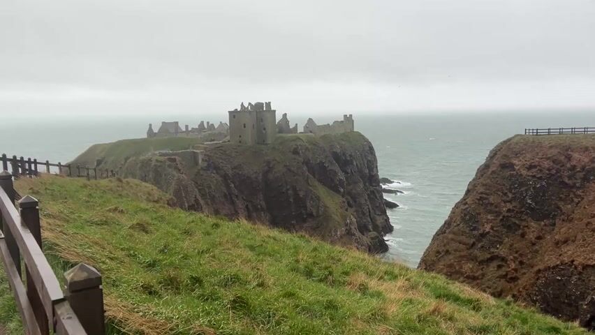 The video presents a view of Dunnottar Castle, a ruined medieval fortress located atop a rocky headland along the coast. The camera slowly zooms in, providing a closer view of the castle's stone structures against a cloudy, overcast sky. The surrounding landscape is characterized by grassy terrain on the adjacent cliff and the ocean below. The waves crash gently against the base of the cliff, adding to the rugged scenery. A wooden fence is visible in the foreground, partially obscuring the view. The general environment suggests a historical, rugged, and natural setting.