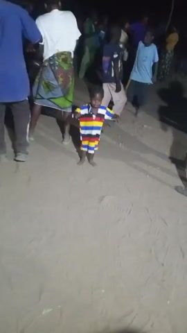 A child stands in the center of the frame, barefoot on a dirt surface. The child is wearing a striped shirt with white, red, yellow, black, and blue stripes. A woman with a patterned skirt and white shirt is partially visible to the left of the child. Two other children are partially visible in the background to the right, one in a blue shirt and tan pants. Other people are visible in the distant background. The child is facing the camera and appears to be standing still, possibly observing something out of frame. A shadow of the child is visible on the dirt surface. There is faint ambient light on the scene. There is music and speech in the audio.