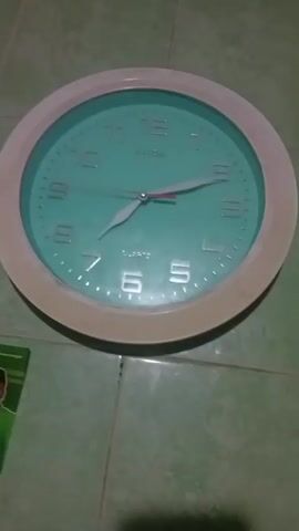 The video shows a close-up of a light-colored wall clock with a teal face. The clock has white Arabic numerals and silver hour and minute hands. The hour hand is pointing between the 10 and 11, and the minute hand is pointing at the 2. The second hand is red and is pointing at the 1. The clock is mounted on a wall with green tiles. There is a shadow on the wall beneath the clock.