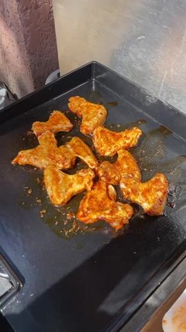 The video shows a close-up of several raw chicken wings coated in a reddish sauce, arranged on a black, flat-top grill. The grill appears to be slightly greasy, and the wings are unevenly spaced. The background includes a textured wall and a stainless-steel surface.