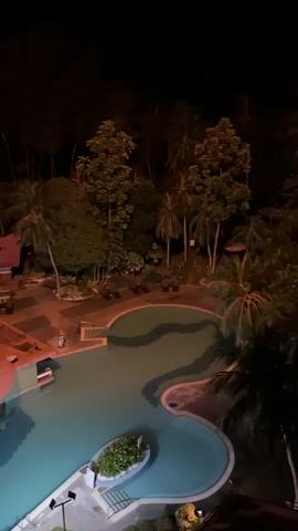 This video showcases an aerial view of a resort's swimming pool area at night. The scene is illuminated by artificial lights, highlighting the turquoise water of the swimming pool and the surrounding tropical landscaping.

The pool features a unique, irregular shape, with a central island housing lush greenery. A prominent feature is a circular, open-air bar or cafe structure within the pool, its orange, conical roof illuminated from below, casting a warm glow. Further into the resort, more buildings with similarly styled orange roofs are visible, along with lounge chairs and pathways.

The backdrop is a dense, dark jungle, characteristic of a tropical location, with numerous palm trees silhouetted against the night sky. The overall ambiance is serene and tranquil, suggesting a peaceful evening at a tropical resort. The location is identified as George Town, Malaysia. There are no people or specific activities explicitly captured in the frames, focusing solely on the architectural and natural elements of the resort at night.