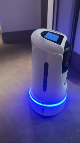 The video shows a white and black robot with a blue light around its base moving down a carpeted hotel hallway. The robot has a screen on top. The hallway has doors on both sides.
