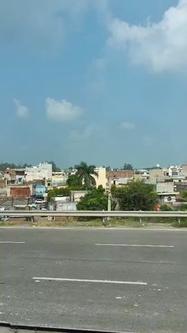 The video shows a highway with a village in the background. The village has a mix of buildings, some with red roofs and others with white or cream-colored walls. There are trees and foliage interspersed throughout the village. The sky is partly cloudy. The camera appears to be moving, suggesting it is mounted on a vehicle traveling on the highway.