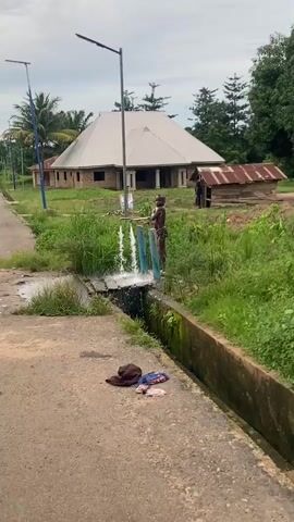A young boy, shirtless and wearing patterned briefs, stands on a wooden plank over an open drainage ditch, his skin covered in soap suds. He holds a bar of soap and washes himself as water flows from a spigot above him, creating two streams that fall into the ditch. In the background, a large building with a light-colored roof is visible, along with a smaller, dilapidated shack. Green foliage and trees surround the area, and another person can be seen in the distance near the shack. On the paved ground in the foreground, a brown hat and some other clothing items are discarded.