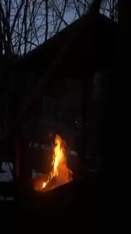 The video depicts a square fire pit with burning wood inside. It is located outdoors, seemingly in a wooded area, as indicated by the bare trees visible in the background. The fire pit has a partial roof or covering above it. The scene is dimly lit, suggesting it's either dusk or nighttime. Towards the end of the video, a hand holding sticks appears near the fire pit, implying someone is tending to the fire.