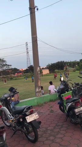 The video shows an outdoor scene with a grassy field where several people are playing soccer. In the foreground, several motorcycles are parked on a red brick-like surface. Some people are sitting on a low green wall along the edge of the field, watching the game. The sky is a light blue, suggesting it is either early morning or late afternoon. A tall utility pole with wires runs vertically through the scene, and in the background, there are trees, buildings, and a distant transmission tower. The overall atmosphere appears to be casual and relaxed, possibly a community gathering or practice session.
