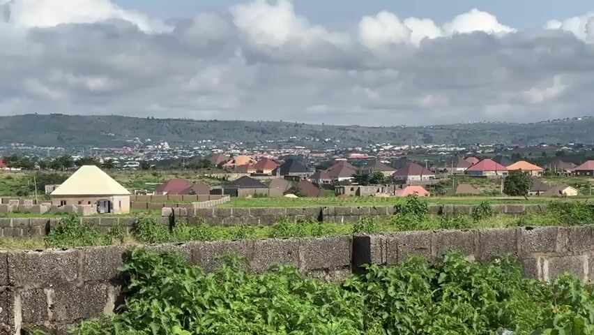 A wide, panning shot reveals a suburban landscape under a cloudy sky. In the foreground, a low wall constructed from concrete blocks is covered in green vegetation. Beyond this, a collection of houses with brightly colored roofs, predominantly red and orange, are spread across rolling hills. The houses appear to be modern and residential. In the distance, more buildings are visible, suggesting a larger town or city, nestled against a backdrop of undulating hills. The overall impression is of a developing residential area in a hilly terrain.