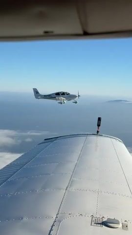 A white Cirrus aircraft with registration N219EL flies through the sky. The view is from another aircraft, showing the wing of the first aircraft in the foreground and the second aircraft flying in the distance. The sky is clear and blue.