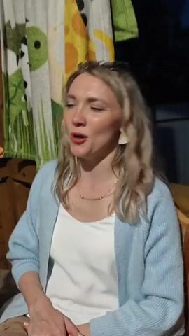 A woman with blonde, wavy hair is sitting on a wooden bench. She is wearing a light blue cardigan over a white top and tan pants. The woman has earrings and a necklace on. She is looking to her right and then turns to look at the camera. She smiles, then places her hand on her chest and makes a gesture with her other hand. She opens her mouth as if to speak or sing and then her hands move again. The background consists of a wooden structure and a curtain with a cartoon pattern featuring giraffes and other animals. The lighting suggests it is evening.