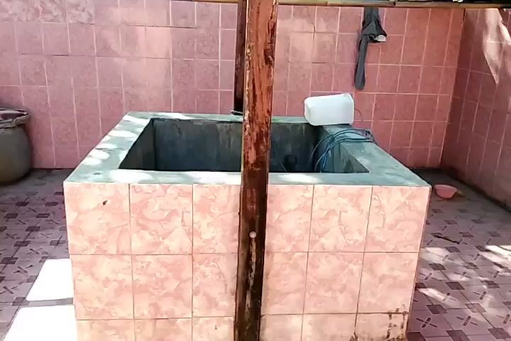 A square-shaped well is the focal point of the video. The well is made of concrete and lined with pink tiles. Inside the well, there is a white plastic container and some electrical cords. Two wooden posts are positioned near the well. The ground around the well is also tiled with pink and white patterns. The walls in the background are tiled with pink tiles, and some shadows from trees are visible on the ground. A gray piece of clothing is hanging on the wall in the upper right corner of the frame. A beige pot is visible on the left side of the frame, near the tiled wall.