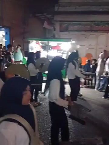 A marching band comprised of young people in white shirts and headscarves is performing in a street at night. They are carrying brass instruments and appear to be moving forward. Several food stalls and shops are visible in the background, with lights illuminating the scene. The overall atmosphere suggests a lively street performance or parade.