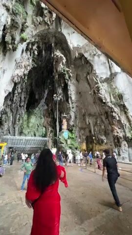 The video depicts the interior of a large cave. A woman in a red dress takes a photo with her cell phone of the cave's interior. The cave's walls are textured with white and gray rock formations, and some green plants grow on the walls. A figure stands on a raised platform, illuminated by a streetlight. Many people are walking and sitting in the large, open space within the cave. The cave is lit by natural light filtering in from the entrance. The floor appears to be wet.