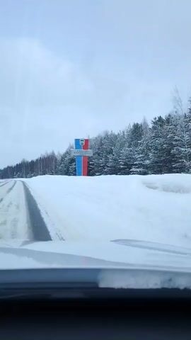 A car is driving on a snow-covered road. The road is mostly clear of snow in the tire tracks, but the shoulders are deep with snow. On the left is a sign in Russian that is red, white, and blue. Behind the sign and to the right of the road is a line of trees. The sky is overcast.
