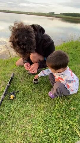 The video shows a man and a child near a lake. The man is on his knees, wearing a black hooded sweatshirt and dark pants. He has dark, curly hair and appears to be working with his hands, possibly preparing fishing gear. The child, who is much shorter, is also on their knees, facing the man and looking at what he is doing. The child is wearing a white long-sleeved shirt with colorful graphics and gray pants with purple shoes. To the left of the man is a fishing rod with a reel attached. The ground is covered in green grass. The lake stretches out behind them, reflecting the sky, which is partly cloudy and appears to be either dawn or dusk. In the distance, across the lake, are some trees and possibly a building.