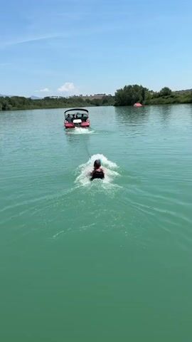 A red boat travels across a bright green body of water on a sunny day, leaving a white wake behind it. A person is water skiing behind the boat, kicking up spray from the water. Trees and hills form the shoreline in the background. The sky is blue with a few white clouds.