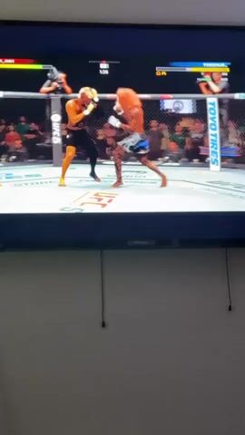 The video shows a screen of a person playing a UFC fighting game. The game is in first-person perspective and shows two fighters in the octagon. One fighter, wearing black shorts, lands a kick that knocks the other fighter, wearing blue shorts with a tiger print, to the ground. The match continues, and they begin to exchange punches. The television the game is being played on is mounted on a light-colored wall. Black cords dangle below the television.  The crowd is visible in the background of the game, and camera operators are present outside of the octagon. There are sponsors' names visible on the canvas.