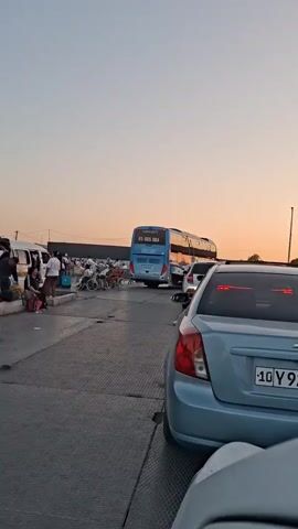 A blue bus with "85 005 DBA" on its back is parked in a lot. Several white vans are parked on the left side of the frame. A woman walks from left to right in the foreground. A man carrying a young boy walks from left to right behind the woman. The boy is resting his head on the man's shoulder and wearing a cap. The man is wearing a white t-shirt with "KAZAKHSTAN" written on the back and tan shorts. The sky is a pale orange, suggesting sunset.