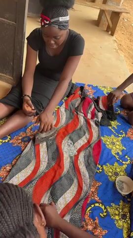A medium shot shows three people gathered around a colorful textile spread on the ground. A young woman in a black dress and headband sits on the ground, pressing an iron onto a red, black, and white dress laid on the textile. The dress has embellishments of small silver beads. Another person, visible from the shoulders down, is seated to the right of the woman. Their hands are picking beads from a small white bowl. The third person is visible from the back of the head, with braided hair. They are also picking beads. The textile on the ground is blue and yellow with floral patterns. The background shows the side of a building with a brown door and a partially visible wooden table. The ground is a mix of dirt and concrete.