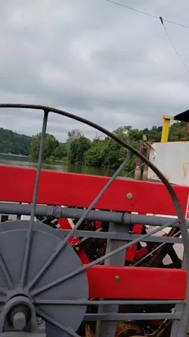The video opens with a close-up of a paddlewheel on a ferry, the red and gray metal blades are visible. A "No Smoking in Vehicles" sign is mounted on the side of the ferry. The camera then shifts to a view through a car's tinted window, showing a white Tesla Model Y parked on the ferry. The Tesla's rear is facing the camera, and its license plate is partially visible. The ferry is surrounded by trees and calm water. The video continues to show the Tesla from different angles as it is on the ferry. Another sign reading "Please Stay in Vehicle" is visible. The ferry appears to be in motion, crossing a body of water. The sky is overcast.