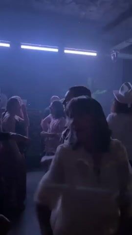 The video depicts a dimly lit dance floor with people dancing and enjoying themselves. The lighting consists of white, blue, and red lights illuminating the space. People are seen with their arms raised, moving to the music, while some are recording on their phones. The room is mostly dark, but a few figures can be seen clearly silhouetted against the lights.