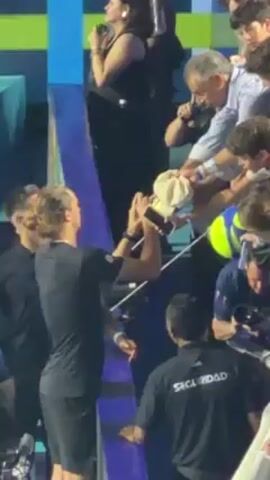 The video shows a man with long blond hair, wearing a black shirt and shorts, standing behind a short blue barrier with a metal railing. He appears to be taking pictures with his phone of something to his right, where several hands are reaching towards him, holding out items like a white hat and tennis balls. Other people are around the barrier, including a woman in black sitting above on some steps, a man in a light blue striped shirt, and another man in a black shirt with the word "SEGURIDAD" on the back. There is also someone wearing a shirt that says “CREW”. The background consists of multi-colored blue and green surfaces. The lighting seems bright, and the scene looks busy.