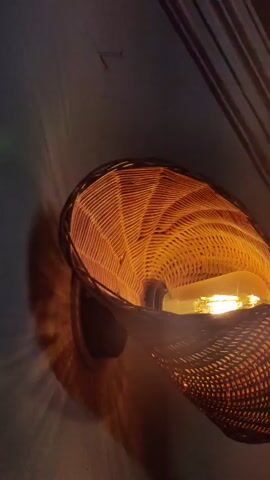The video shows a close-up, slightly shaky shot of a woven basket-like light fixture mounted on a wall. The basket is illuminated from within by a warm, yellow light, likely from a candle or a low-wattage bulb. The light casts a patterned shadow on the wall behind it, creating a dynamic visual effect. The camera angle is from below and to the side, emphasizing the texture and translucency of the woven material. The overall atmosphere is warm and dimly lit, suggesting a cozy or ambient setting.