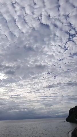 This video is an aerial shot of a calm sea under a cloudy sky. The clouds are textured, appearing in waves and patterns, with some patches of sunlight breaking through. In the distance, a small red kayak with two people is visible on the water. The horizon shows a faint outline of land on the left side of the frame. The camera angle is from above, looking down at the seascape.
