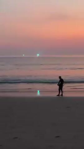 The video shows a beach scene at sunset. The sky is a gradient of pink, orange, and light purple. A man in dark clothing stands on the wet sand near the water's edge, looking down. The waves are gentle and reflecting the colors of the sky. In the distance, several people are wading in the water. There are also two bright green lights visible far out on the water.