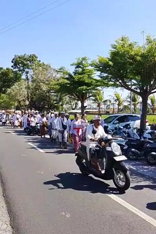 A group of people are walking in a procession along a paved road. Most of the people are wearing white traditional clothing. A person on a scooter is riding past the procession. The sky is clear and blue, and there are trees on the side of the road. The time stamp is 00:00.