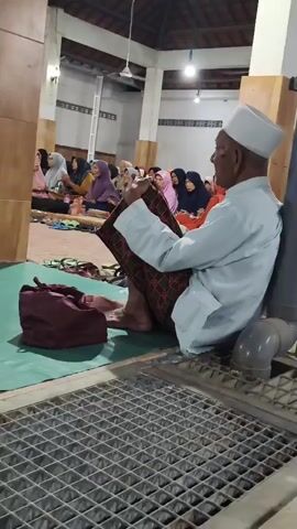 A group of people are gathered inside what appears to be a mosque or prayer hall. In the foreground, an elderly man wearing a white cap and a light blue shirt sits on the floor, leaning against a pillar. He is wearing a sarong with a red and green patterned design. To his left is a dark red bag. A metal grate is visible in the bottom of the frame. In the background, several women are seated on the floor, some wearing hijabs in various colors. They appear to be engaged in prayer or listening to an address. The room has a high ceiling with exposed wooden beams and white walls. Lighting fixtures are visible on the ceiling.