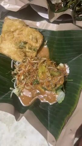 A close-up shot shows two plates of food. The plate in the foreground features a fried item topped with a peanut sauce and some vegetables. The plate in the background contains rice, meat, vegetables, and a dollop of red sauce. The food is served on banana leaves and wrapped in paper. The lighting is dim, suggesting an indoor setting.