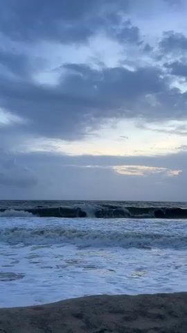 The video shows a choppy ocean under a cloudy sky. Waves crash on the shore, creating foam. The lighting suggests it is either dawn or dusk. The camera is focused on the ocean and the sky, with a sliver of the beach visible in the foreground.
