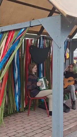 The video shows an outdoor setting with a covered shelter decorated with colorful streamers. A young person with glasses is playing an accordion while sitting on a red chair. Another person with green-tinted hair is playing an acoustic guitar. They are both seated under the shelter. In the background, a building with wooden siding is visible, adorned with colorful triangular flags. There is a bicycle with flowers in the background. The ground is paved with bricks. The video captures a musical performance or gathering.