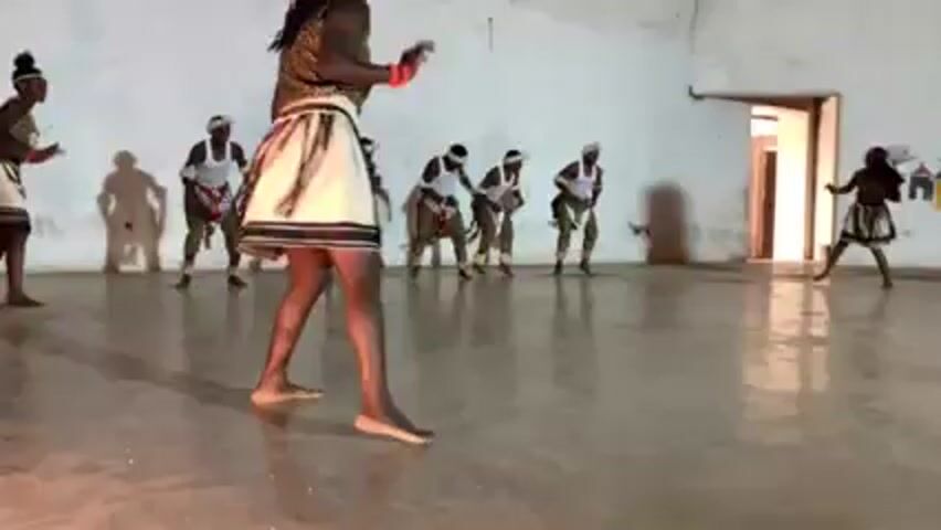 A group of eight individuals are engaged in a choreographed dance within a large, sparsely decorated room. The dancers, dressed in similar attire consisting of light-colored skirts with dark vertical stripes, leopard print vests, and white headbands, move in unison, bending at the waist and stepping forward. The women wear their hair in high buns, and at least one wears red armbands. The room has white walls, a light-colored floor, and minimal decor, though a mural featuring simple drawings of houses and a tree can be seen on one wall. A doorway is visible on the right side of the frame, leading to an adjacent room. The lighting is subdued, casting shadows of the dancers on the walls and floor, which reflect the light.