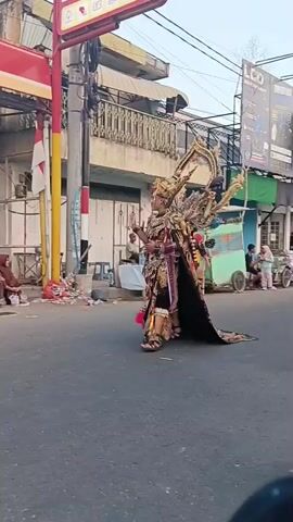 A person in an elaborate gold and blue costume walks down a street. The costume features large, ornate wing-like structures on the back and a detailed headdress. The person holds a staff. Spectators, including several women wearing hijabs and some men and children, are seated on the side of the road, observing the parade. Some people are standing in the background near shops and stalls. There is trash on the ground near the spectators. A red and yellow sign is visible on a pole. The sky is visible and appears to be daytime. The scene appears to be a cultural event or parade taking place on a street in an urban or semi-urban setting.