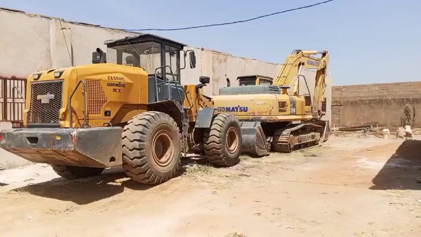 The video shows a yellow Lonking ZL50HC wheel loader and a yellow Komatsu PC360 excavator parked next to each other in an outdoor area. The excavator is a tracked vehicle, while the Lonking has large, thick tires.  The vehicles are parked in a yard with a dirt surface, and there are several buildings in the background. The sky is clear. A few other items are in the environment, including a table and chair. The video is a static shot, and no one is visible.