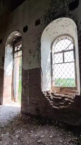 The video shows the interior of a ruined building. The walls are mostly brick with patches of white paint peeling off. There are multiple arched windows and doorways, some with open frames and others covered with metal lattice. The floor is covered in debris. The ceiling is arched and heavily stained with dark brown and black patches. Light streams in from the windows, illuminating the dusty interior.