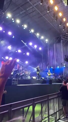 The video shows a live concert performance on an outdoor stage. Two male musicians are visible on stage, both playing guitars. One man in a dark shirt and jeans stands center-stage, while another man in a lighter shirt stands to his right. A drum set is visible behind them. The stage is lit with bright purple and white lights, and there is a considerable amount of smoke or haze in the air. A crowd of spectators is partially visible in the foreground, behind a railing. The shot is taken from a raised vantage point, looking down towards the stage.