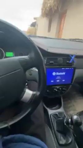 A person is sitting in the driver's seat of a car. The car's dashboard is visible, showing a Bluetooth screen. The steering wheel is black with silver accents and has a Chevrolet logo in the center. The person is wearing a blue jacket. The car appears to be parked in front of a building with a straw roof. The interior of the car is visible, including the gear shift, dashboard, and seats. The car's instrument panel is visible through the steering wheel. The person's hand is visible on the steering wheel.