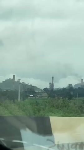 The video shows a wide shot of an industrial landscape under a cloudy sky. In the foreground, a portion of a vehicle's interior is visible, including a dashboard with a black and white striped pattern and some writing. In the midground, there is lush green vegetation and trees surrounding a cluster of industrial buildings. Tall smokestacks are prominent, with plumes of smoke rising into the air, indicating active industrial activity. There are also some smaller buildings and structures scattered among the trees. In the background, rolling hills are covered in green foliage. A small airplane is visible in the sky. The overall atmosphere is overcast and industrial.