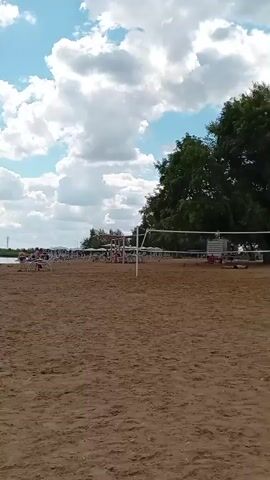 The video begins with a wide shot of a sandy beach next to a body of water under a bright, cloudy sky. In the foreground, the sand is mostly empty, with some scattered beach chairs and a volleyball net visible. As the video progresses, the camera pans left, revealing more of the beach and the water. Several people can be seen on the beach and in the water, some swimming or playing near the shore. The background shows a line of trees along the far bank of the water. The camera continues to pan, offering a broader view of the scene, showing more beachgoers and the expansive water. The video concludes with a clear view of the water with people enjoying themselves.