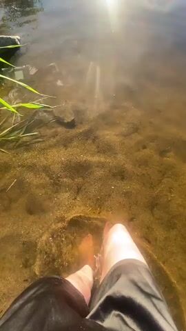 The media file captures a person's bare legs and feet submerged in shallow, clear water. The scene is set at the edge of a body of water, possibly a lake or river, as indicated by the presence of reeds and rocks along the bank. The sun is shining brightly, creating a warm, golden glow on the water's surface and reflecting in a prominent lens flare in the upper right portion of the frame. The bottom of the water is sandy and appears to have small pebbles. The person is wearing dark-colored shorts. They are gently moving their legs, causing ripples and splashes in the water. The activity appears to be a leisurely moment of cooling off in the water on a sunny day. The overall mood is peaceful and relaxed. There is no visible text in the media file.