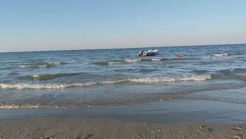 The video shows a calm beach scene with gentle waves rolling onto the sandy shore. The sky is clear and blue, indicating fair weather. In the distance, a small white boat with an outboard motor is anchored in the water. Several orange buoys are also visible, marking an area in the sea. The water is a deep blue, reflecting the clear sky. The sand on the beach is a light color, with some scattered shells visible. The scene is tranquil and peaceful, suggesting a pleasant day at the beach.