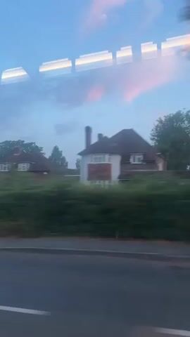 The video shows a blurred, motion-effected view from a window, likely a vehicle, looking out at houses and greenery. The sky is a soft blue with streaks of pink and orange clouds, suggesting either dawn or dusk. The houses appear to be residential, with hedges and trees in front of them. The road in the foreground is also blurred, indicating movement. Reflections of rectangular lights are visible on the window pane at the top of the frame.