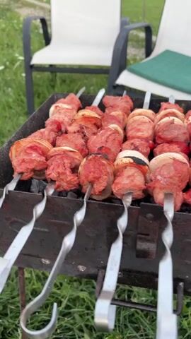The video shows a close-up of several skewers of meat and onions cooking on a barbecue grill. The skewers are made of metal and are placed horizontally over hot coals. The meat appears to be raw or lightly cooked, and some pieces have red onions interspersed between them. The grill is dark metal, and some embers are visible underneath the skewers. In the background, two outdoor chairs with white canvas seats and black frames are visible, with one chair having a green cushion on it. The chairs are positioned on a grassy area. The overall setting appears to be outdoors, possibly a backyard or patio, with green grass and some foliage in the background. The lighting suggests it might be daytime or early evening.