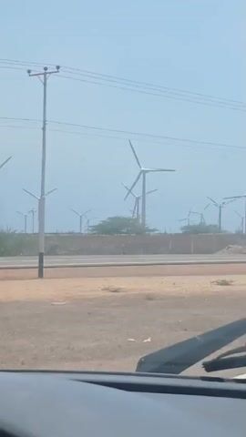 The view from inside a car shows a field of wind turbines. The turbines are white and have three blades, and they are arrayed in rows behind a concrete barrier along a road. The landscape is dry and dusty, with only a few small trees and shrubs. The sky is light blue and slightly hazy. Power lines run overhead. The windshield wipers of the car are visible at the bottom of the frame. The car is moving, as evidenced by the changing perspective of the scene.
