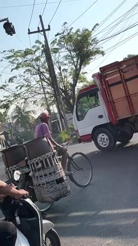 A video shows a man riding a black scooter. He is wearing a dark vest over a green shirt and black pants. He is looking ahead. Behind him, a large orange truck is visible. A second person is partially visible on the back of a motorcycle parked next to the truck. The scooter has a license plate with the numbers 3487 WDE. Overhead, power lines crisscross the scene. The background shows trees and buildings, suggesting an outdoor, urban or semi-urban environment. The time stamp at the beginning of the video reads 00:00.