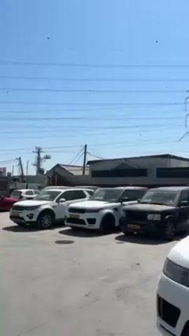 Here is a description of the video.

The video shows a row of Range Rovers parked in an outdoor lot. The cars are mostly white and black, and some of them have their hoods open. There is a large metal building in the background with open garage doors. The sky is blue and clear. The scene appears to be a car dealership or repair shop.